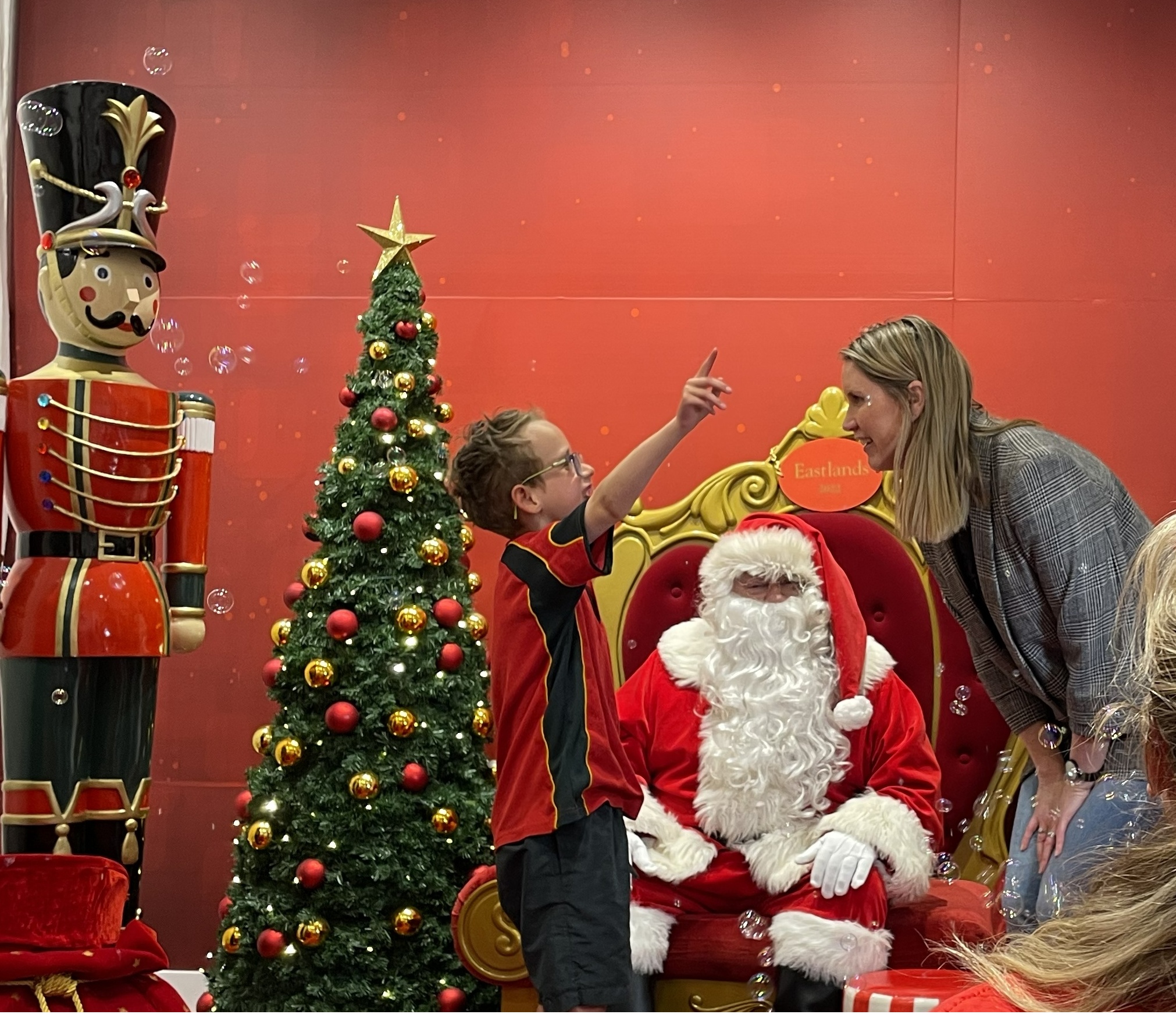 7yo boy with glasses standing in front of Santa claus happily points at his mother. Beside him is a fully decorated christmas tree and a tall wooden toy soldier